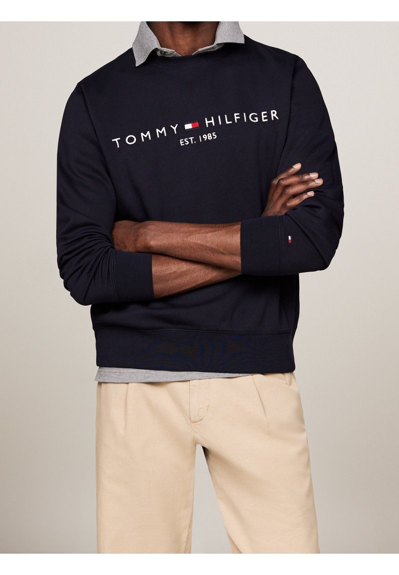 Uomo che indossa una felpa navy di Tommy Hilfiger sopra una camicia grigia con pantaloni beige, braccia incrociate in vita.