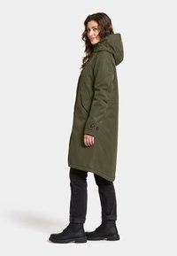 Olijfgroene waterdichte jas met capuchon, lange pasvorm, twee zijzakken en knoopaccenten, gedragen over donkere jeans en zwarte laarzen.