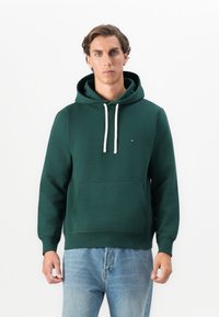 Man with short brown hair wearing a dark green hoodie with white drawstrings and light blue jeans, standing against a plain white background.