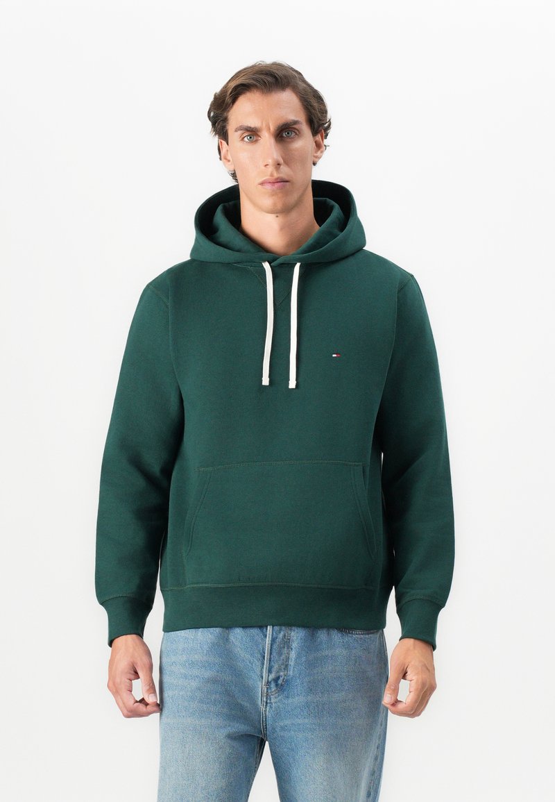 Man with short brown hair wearing a dark green hoodie with white drawstrings and light blue jeans, standing against a plain white background.