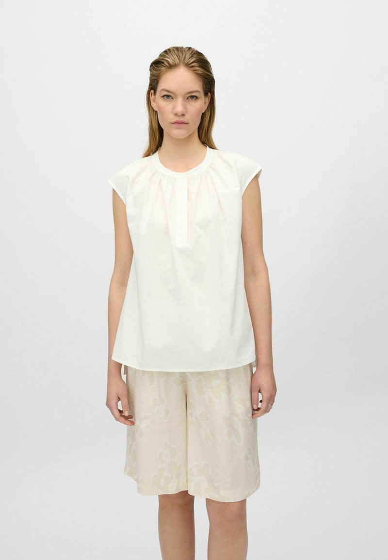 Blusa di cotone bianca con maniche corte a cappuccio, scollo rotondo e dettaglio arricciato davanti, abbinata a pantaloni corti floreali beige chiaro.
