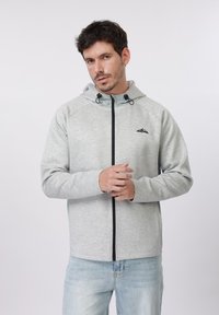 Άνδρας με κοντά σκούρα μαλλιά που φοράει ανοιχτό γκρι φούτερ με φερμουάρ και ανοιχτό μπλε τζιν, στέκεται μπροστά σε απλό λευκό φόντο.