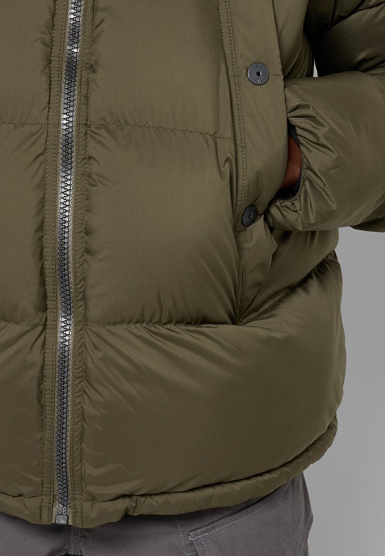 Olivgrüne Pufferjacke mit einem Reißverschluss an der Vorderseite, Druckknopfverschlüssen und elastischem Saum. Glatte, gepolsterte Textur mit diskreten Seitentaschen.