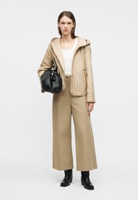 Femme portant une veste à capuche beige, haut blanc, pantalon beige à jambes larges, bottes noires, tenant un sac à main noir, debout devant un fond blanc.