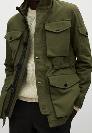 Mężczyzna noszący oliwkową kurtkę z wieloma kieszeniami z klapkami na białym dzianinowym swetrze i czarnych spodniach.