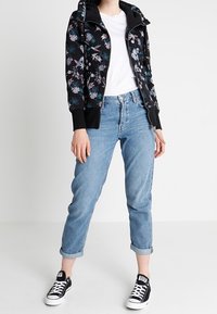 Zwarte ritssluiting hoodie met bloemenborduursels, gecombineerd met lichtblauwe jeans met opgerolde zoom en zwarte sneakers. Simpel wit T-shirt eronder.