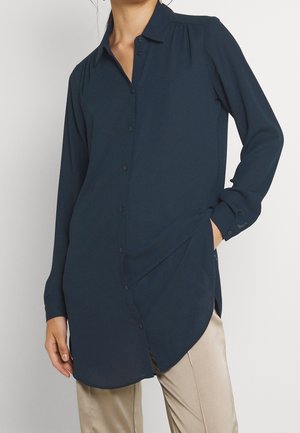 Kvinde iført en lang marineblå skjorte med knapper og lange ærmer og beige bukser, med den ene hånd i lommen, mod en ensfarvet baggrund.