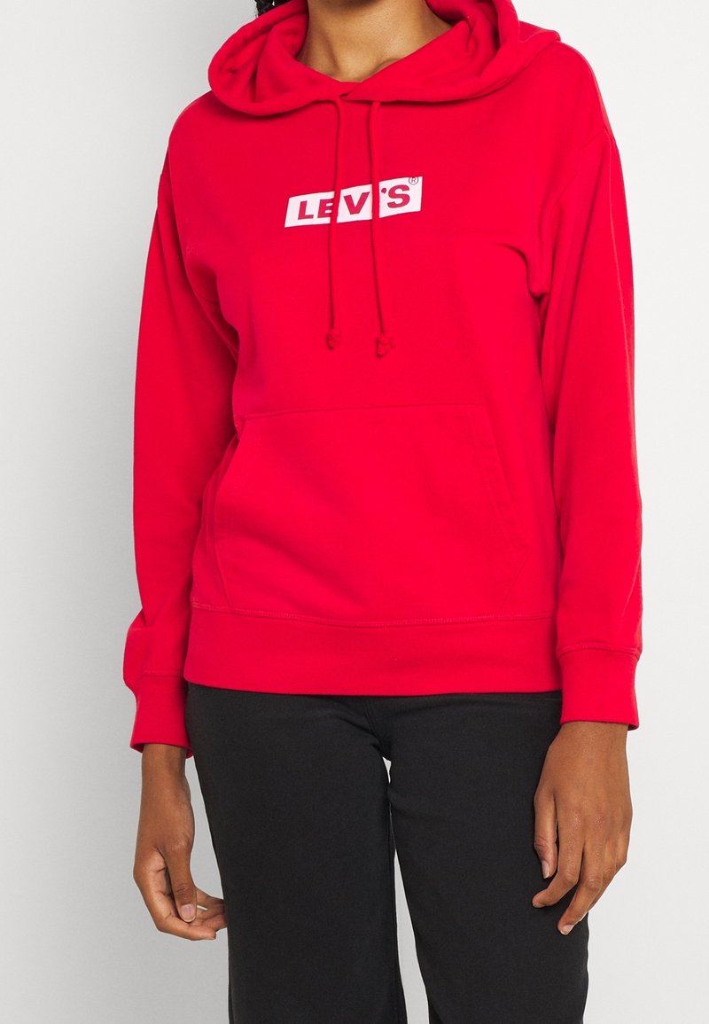 Personne portant un sweat à capuche rouge Levi's avec une poche avant et un pantalon noir, visible du cou aux hanches sur un fond uni.