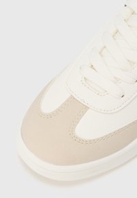 Pantof sport bej și alb, cu partea superioară din piele texturată, vârf rotund, șireturi plate albe și accente subtile de cusături.