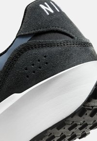 Zapatillas de ante negro con una suela de goma blanca, que cuentan con una marca bordada, perforaciones para transpirabilidad y detalles texturizados.