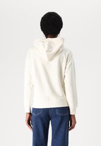 Kremowa bluza z regulowanym kapturem, ściągaczami na rękawach i luźnym krojem, w zestawie z niebieskimi dżinsami. Gładka tekstura materiału.