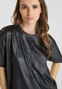 Donna con lunghi capelli castani che indossa un top nero lucido, corto e comodo, su uno sfondo semplice e chiaro.