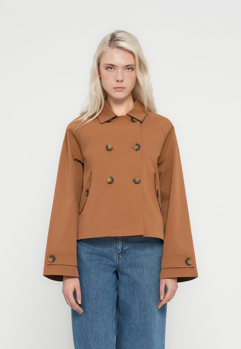 Νεαρή γυναίκα με μακριά ξανθά μαλλιά, ντυμένη με καφέ διπλόφρακο σακάκι με φαρδιά μανίκια και μπλε τζιν, στέκεται μπροστά σε λευκό φόντο.