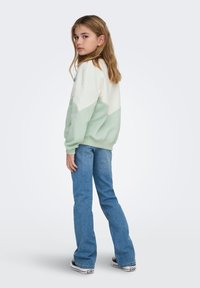 Φούτερ με χρωματική διαμόρφωση σε μίντ και κρεμ αποχρώσεις με άνετη εφαρμογή, συνδυασμένο με ανοιχτό μπλε τζιν καμπάνα και μαύρα-άσπρα αθλητικά παπούτσια.