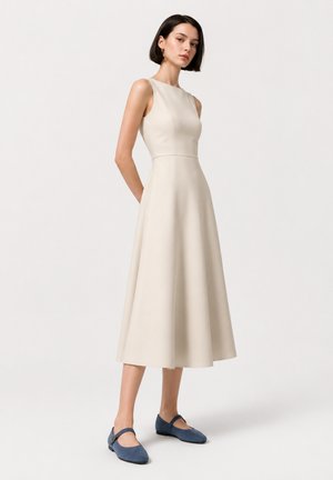 Femme aux cheveux courts et foncés portant une robe midi beige sans manches et des chaussures plates bleues Mary Jane, debout devant un fond clair uni.