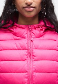 Λαμπερό ροζ γιλέκο με φερμουάρ μπροστά και ρυθμιζόμενο κουκούλα. Διαθέτει υφή με ραφές και κορδόνια στον λαιμό για άνετη εφαρμογή.