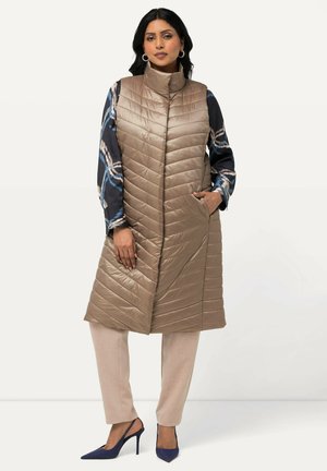 Chaleco acolchado de color marrón claro con cuello alto y bolsillos laterales, llevado sobre una blusa azul con estampado y combinado con pantalones beige y tacones azul marino.