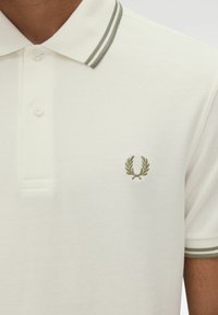 Camisa polo blanca con tejido texturizado, presenta un cuello de dos botones y detalles en verde a rayas. Logotipo bordado en verde en el pecho.