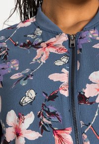 Giacca con zip a tema floreale, caratterizzata da uno sfondo blu con fiori rosa, viola e bianchi, oltre a farfalle. Tessuto testurizzato e colletto a costine.