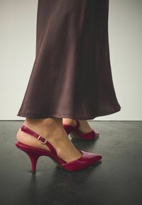 Escarpins slingbacks en cuir verni rouge avec bouts pointus et talon aiguille de 5 cm. Détail de boucle sur la bride de cheville. Jupe fluide marron en arrière-plan.