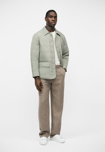 Homme portant une veste matelassée vert clair, une chemise blanche, un pantalon en laine beige et des baskets blanches, se tenant devant un fond blanc uni.