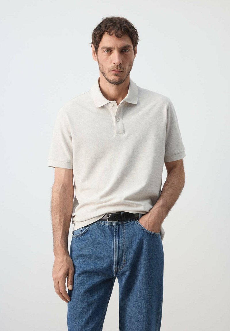 Homme aux cheveux bouclés portant un polo beige et un jean bleu, debout avec une main dans la poche devant un fond uni.