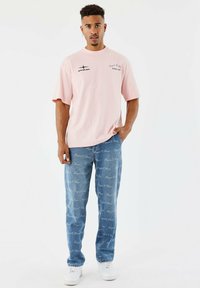 Joven sosteniendo un pie en un fondo blanco, vistiendo una camiseta gráfica rosa clara, jeans azules con escritura blanca y zapatillas blancas.