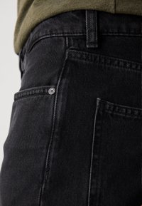 Gros plan d'une personne portant un jean en denim noir et une chemise vert olive, mettant l'accent sur la poche arrière et la zone de la taille.