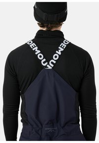 Ski bib overall in donker marineblauw, met verstelbare zwarte banden met witte tekst, gecombineerd met een aansluitend zwart longsleeve basislaag.