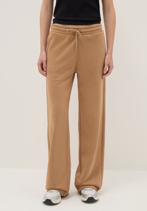Personne portant un pantalon de jogging large couleur beige avec une taille élastique à cordon et des baskets blanches, debout devant un fond clair uni.