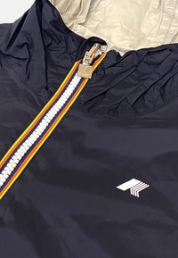 Veste bleu marine avec une fermeture éclair rayée blanche, orange et jaune, une tirette de fermeture éclair argentée, une doublure intérieure beige et un logo géométrique blanc sur la poitrine.