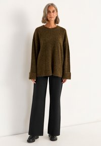 Übergroßer olivgrüner gestrickter Pullover mit weiten Ärmeln und gerippten Bündchen, kombiniert mit schwarzen weit geschnittenen Hosen und klobigen schwarzen Schuhen.