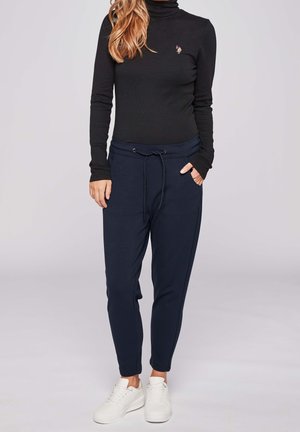 Kobieta w czarnym golfie z długim rękawem, granatowych spodniach dresowych ze ściągaczem i białych sportowych butach, stojąca na tle jasnoszarym.