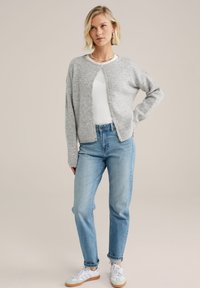 Světle šedý pletený cardigan s kulatým výstřihem, zapínání na knoflíky, spojeno se světle modrými vysokými džíny a bílými teniskami.