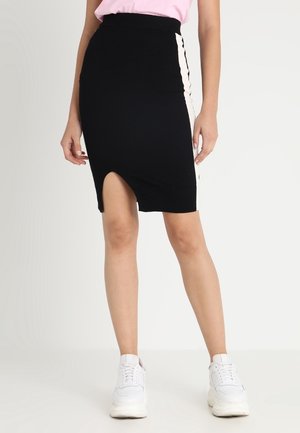 Femme portant une jupe noire longueur genou avec une fente à l'avant et des bandes blanches sur les côtés, associée à des baskets blanches et un haut rose.