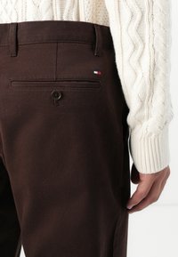 Kleine bruine katoenen broek met een achterzak met een enkele knop en een klein rood, wit en blauw logo patch. Gepaard met een romige kabelgebreide trui.