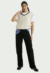 Donna con capelli intrecciati che indossa un maglione senza maniche a treccia bianca sopra una maglietta bianca, pantaloni neri e scarpe da ginnastica nere con strisce bianche.