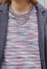 Maglietta a righe a maniche corte in colori pastello; catena in metallo argentato attorno al collo; blazer scuro texturizzato a sinistra.