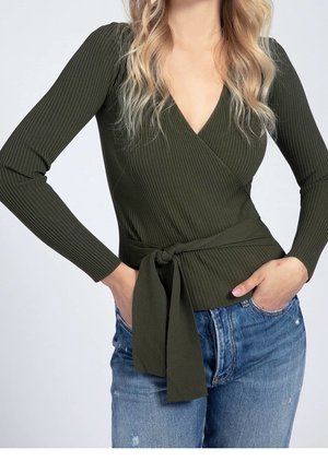 Kobieta z długimi blond włosami, ubrana w przylegający oliwkowozielony prążkowany sweter kopertowy związany w talii oraz niebieskie dżinsy, z rękami w kieszeniach.