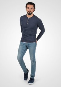 Marineblå langermet henley skjorte med knappdetaljer, kombinert med lyseblå jeans og mørkeblå sneakers. Bakgrunnen er ensfarget og lys.