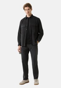  Czarna koszula zapinana na guziki z dwoma kieszeniami na piersi, noszona na czarnej koszulce, w połączeniu z ciemnymi spodniami i jasnobeżowymi sneakersami. Gładka faktura tkaniny.