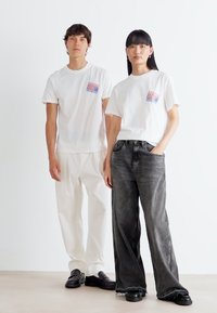 White cotton t-shirts with a colorful graphic patch, paired with loose-fitting white pants and dark gray wide-leg jeans. Both wear black shoes.