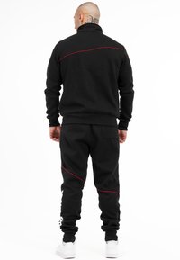 Zwarte trainingsoutfit gemaakt van zachte stof, met een hoge kraag, rode accentstiksels en smalle manchetten en enkels. Witte sneakers maken de look af.