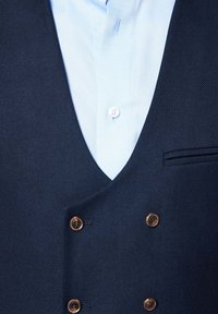 Granatowa, teksturowana kamizelka z głębokim dekoltem w kształcie litery V, zdobiona złoto-brązowymi guzikami, noszona na jasnoniebieskiej koszuli.