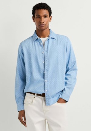 Mann trägt hellblau gemustertes Hemd über weißem T-Shirt und weißen Hosen mit braunem Gürtel, steht mit einer Hand in der Hosentasche.