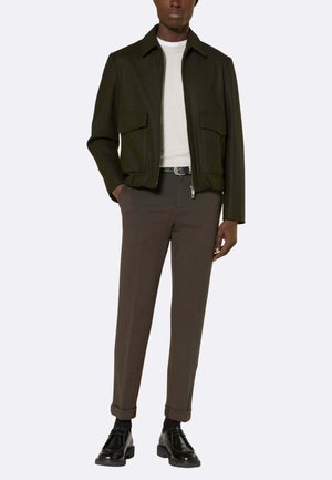 Giacca verde scuro con chiusura a zip e due tasche frontali, abbinata a camicia grigio chiaro e pantaloni marroni. Scarpe nere completano il look.