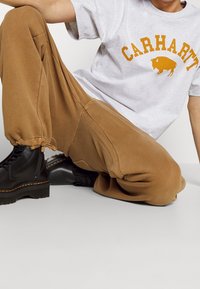 Grå t-shirt med orange "Carhartt" tekst, parret med brune sweatpants og sorte snørebåndsstøvler med en tykk sål og gul syning.