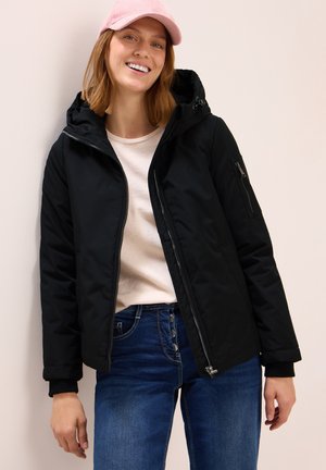Veste rembourrée noire avec une capuche, manches longues, fermeture éclair à l'avant et poches latérales. Portée sur un haut léger et un jean bleu. Casquette rose sur la tête.