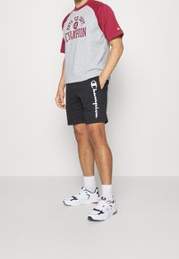 Camisa de manga corta gris y marrón con estampado de "Champion", combinada con pantalones cortos negros que tienen un logo blanco, y zapatillas blancas con detalles.