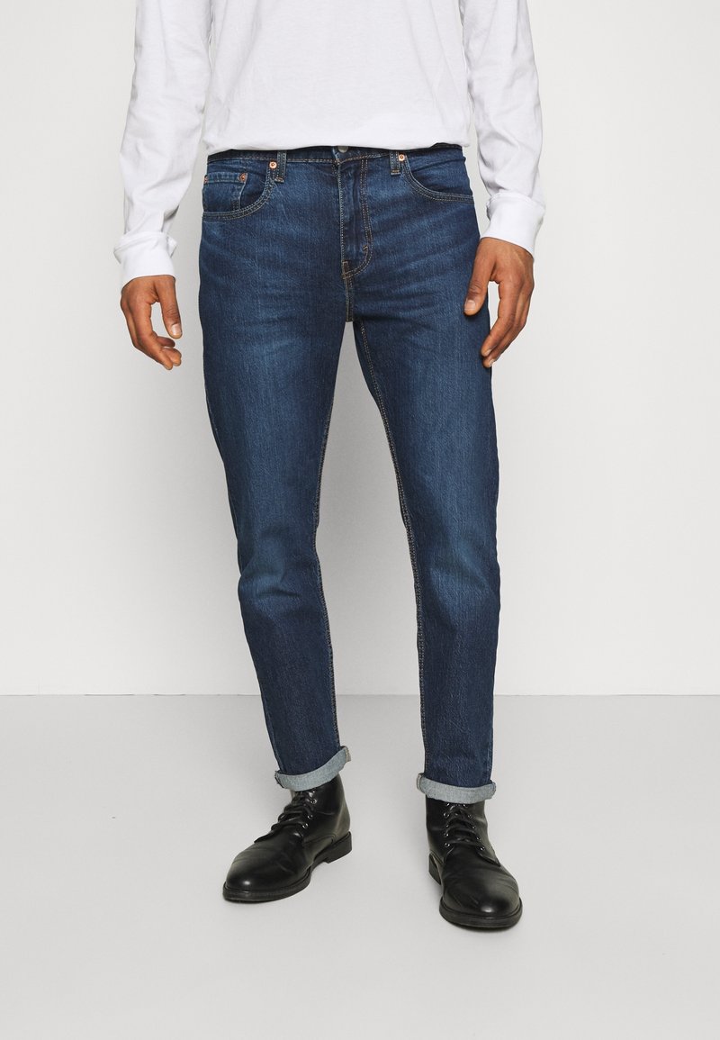 Homme portant un jean bleu foncé retroussé, des bottes noires à lacets et une chemise blanche à manches longues, debout devant un fond uni.